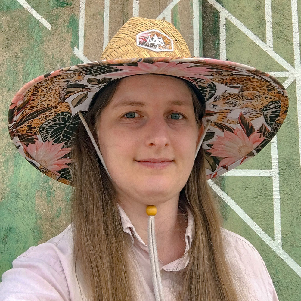A headshot portrait of Ashley. She ha long straight hair, blue eyes and is smiling slightly. She is wearing a big straw sun hat with a floral design under the brim, and a light pink button down shirt. Behind her is a textired green wall with a mural of sharply painted abstract white geometric lines partially visible.